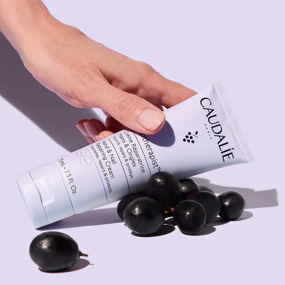 Hand holding Caudalie hand cream tube with black grapes on a light purple background.