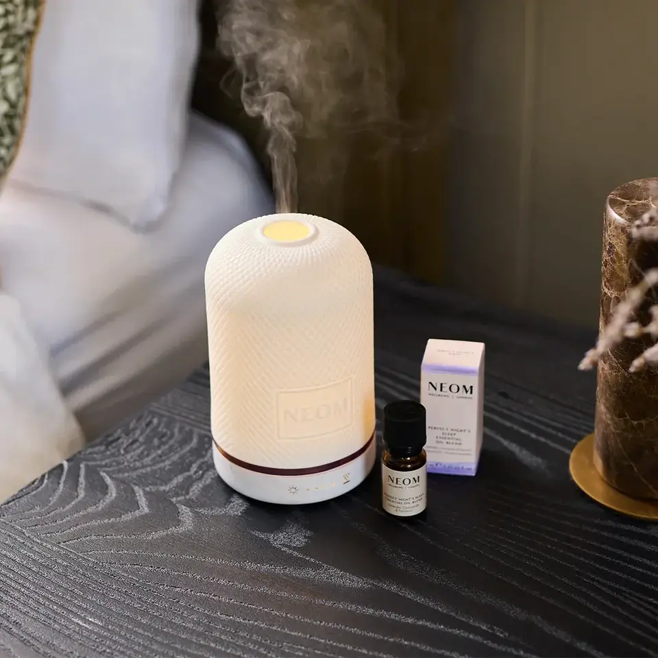 White diffuser emitting mist, with NEOM essential oil and box on a dark wooden table.