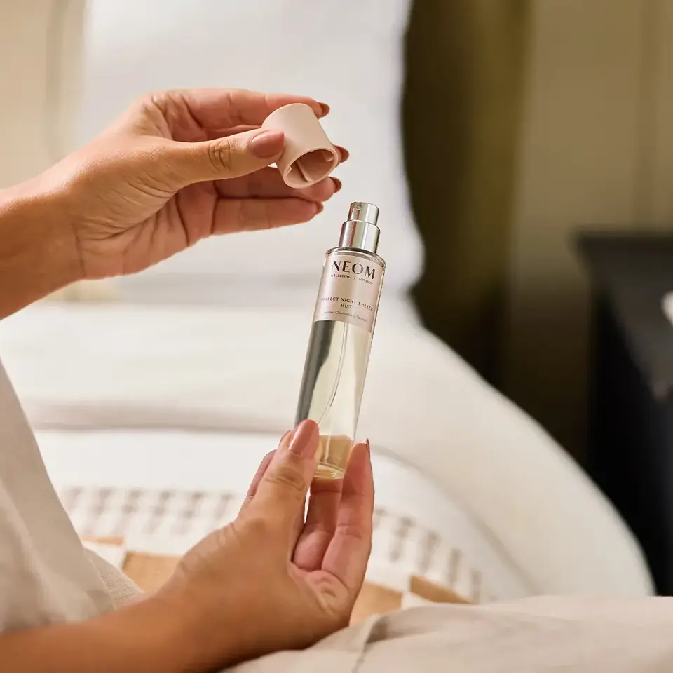 Hands holding a NEOM sleep spray bottle with cap removed, near a bed.