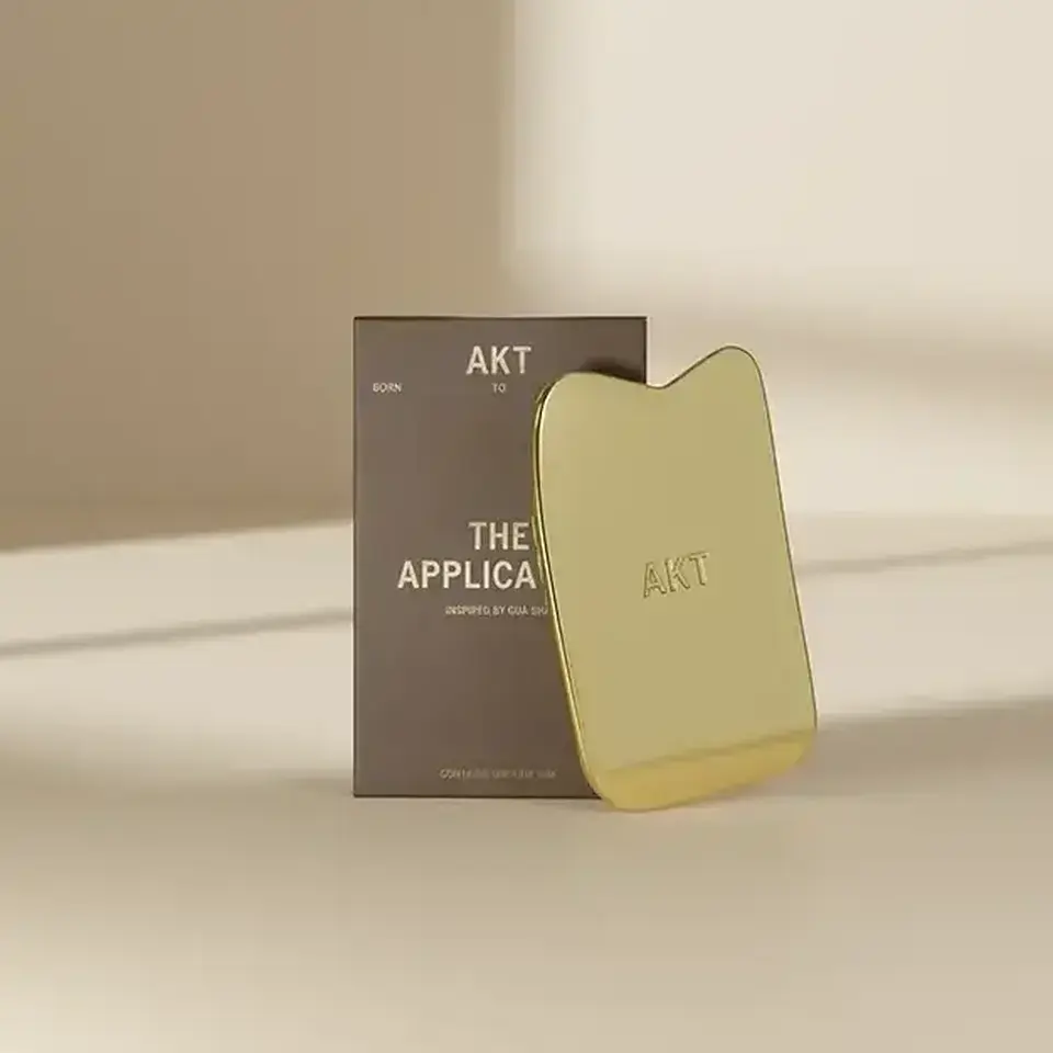 Gold AKT applicator with packaging on a beige surface.