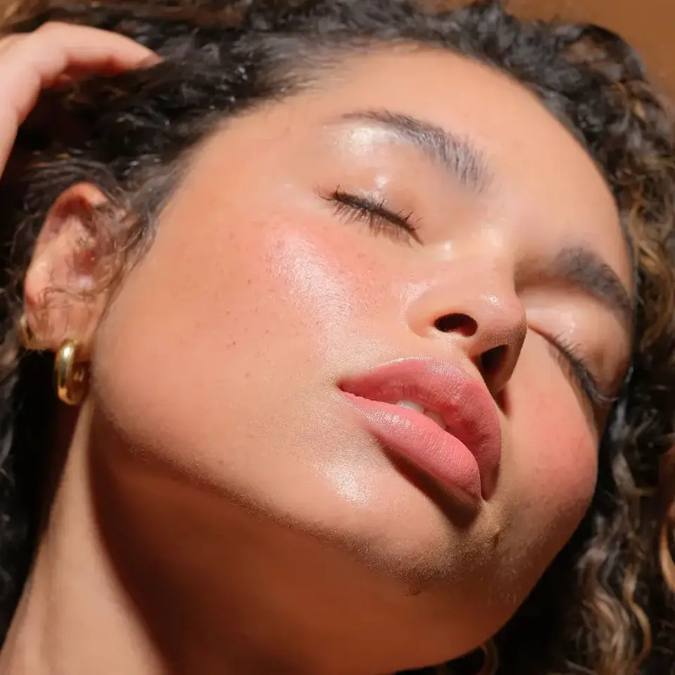 Person with glowing skin, eyes closed, wearing gold hoop earrings. Text: "Living Luminizer".