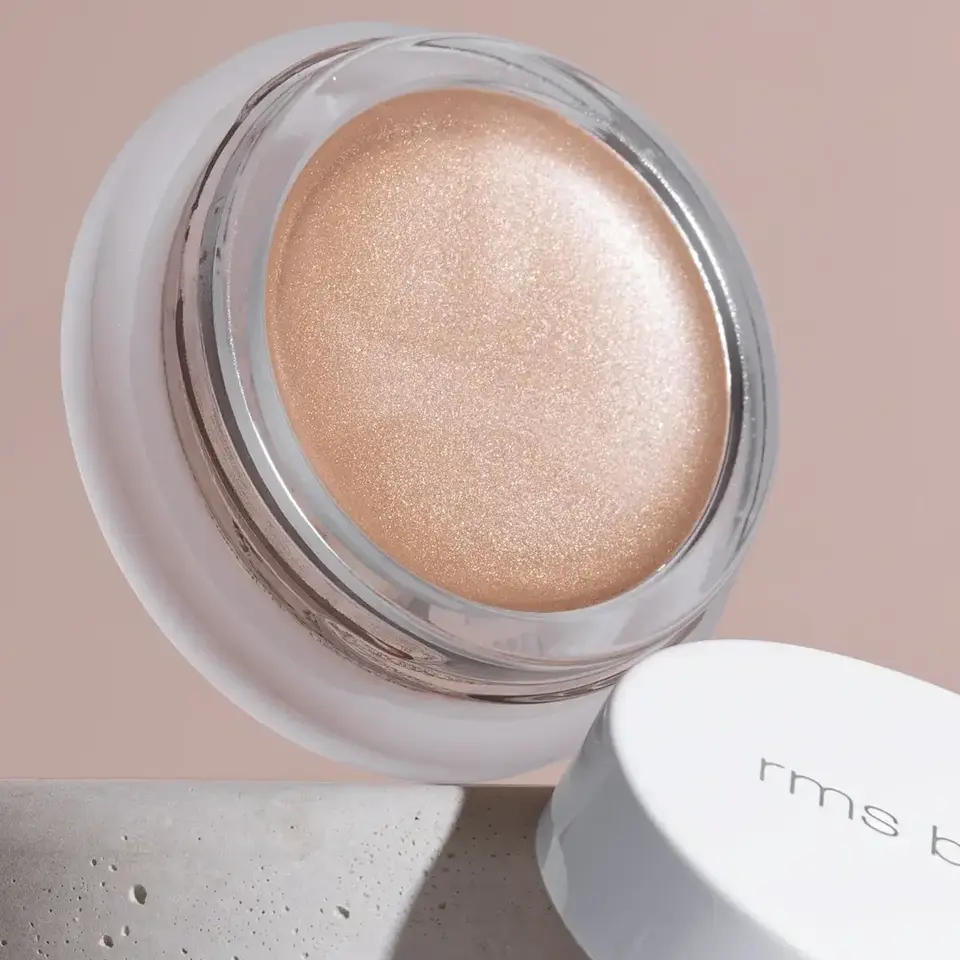 Open jar of shimmering peach cream with "rms beauty" on the white lid, set on a stone surface.