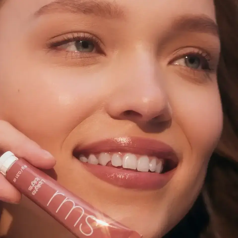 Smiling person holding RMS Beauty lip gloss tube. Text: "RUMOR".