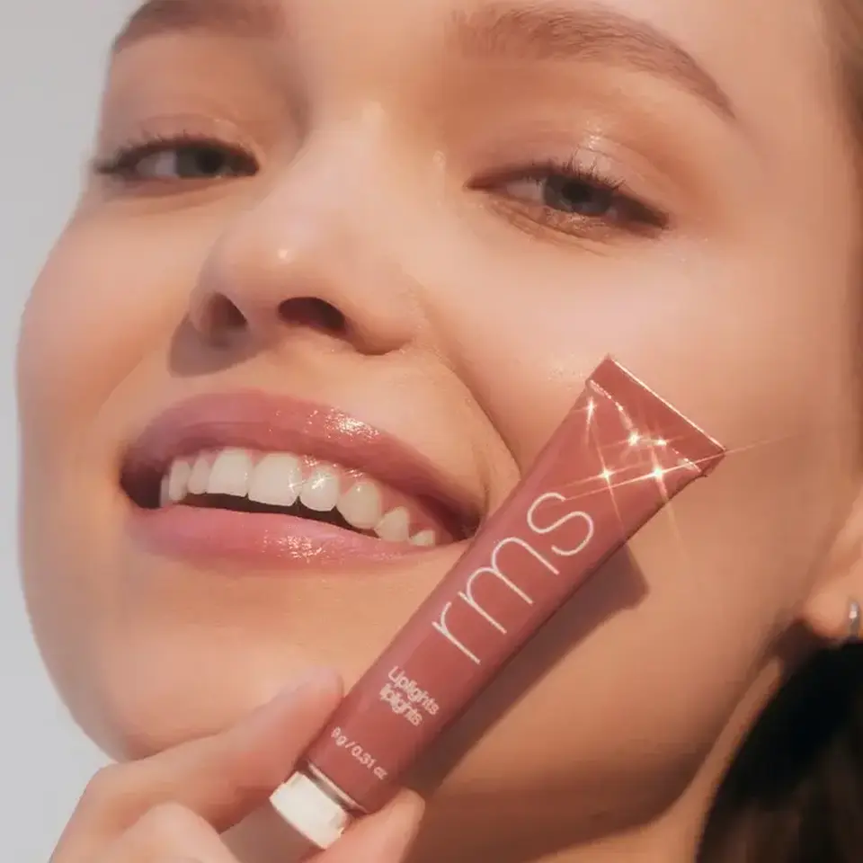 Smiling person holding a tube of RMS Beauty lip gloss labelled "Crush".