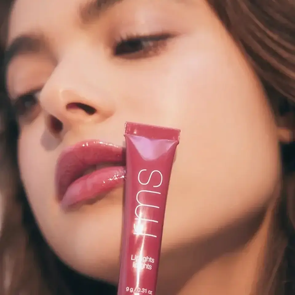 Woman holding a pink lip gloss tube labelled "SUU" near her face.