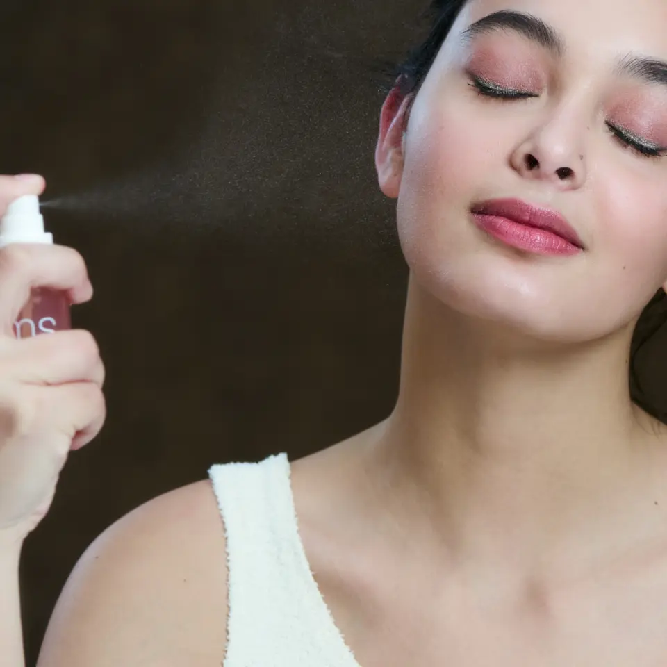 Woman spraying facial mist with eyes closed, wearing a white top.