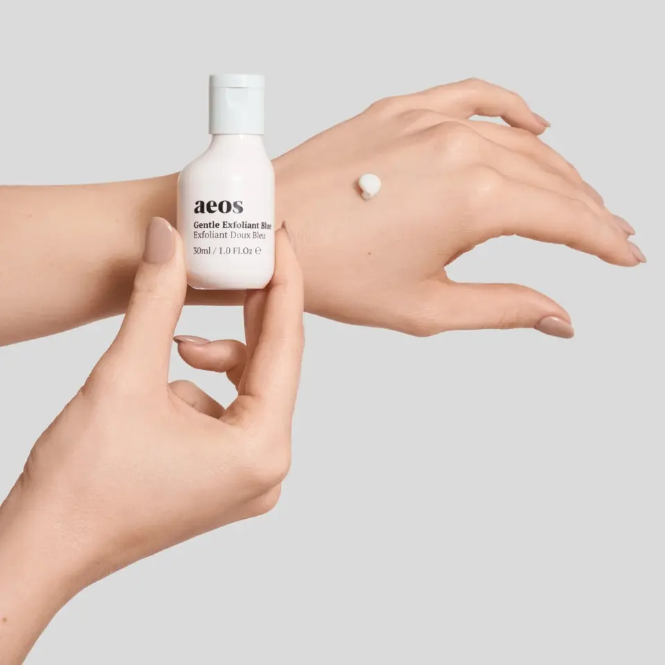 Hands holding aeos Gentle Exfoliant Blue bottle, cream on back of hand.