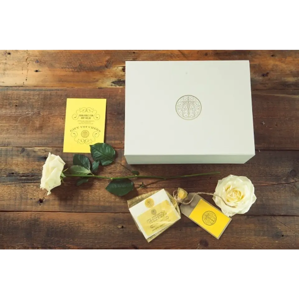 White gift box with logo, yellow card, soap, and white roses on wooden table.