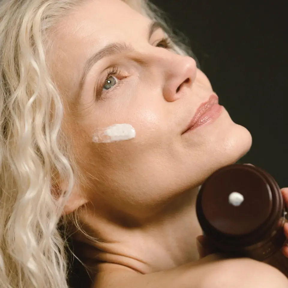 Woman applying cream to cheek, holding brown jar.