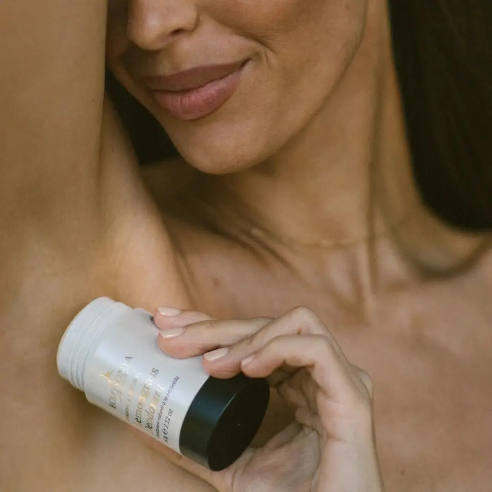 Woman applying deodorant to underarm, holding a white container with black cap.