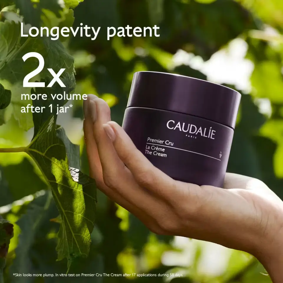 Hand holding Caudalie Premier Cru cream jar, green leaves in background. Text: "Longevity patent, 2x more volume after 1 j...