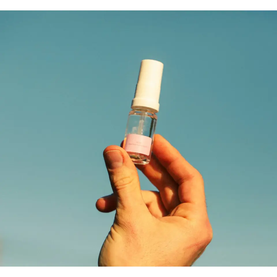 Hand holding a small spray bottle with a pink label against a clear blue sky.