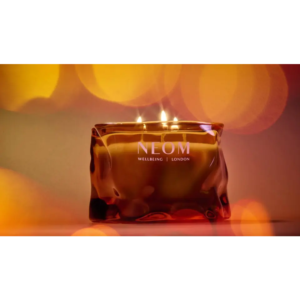 Amber glass candle with "NEOM Wellbeing London" text, lit with three flames.
