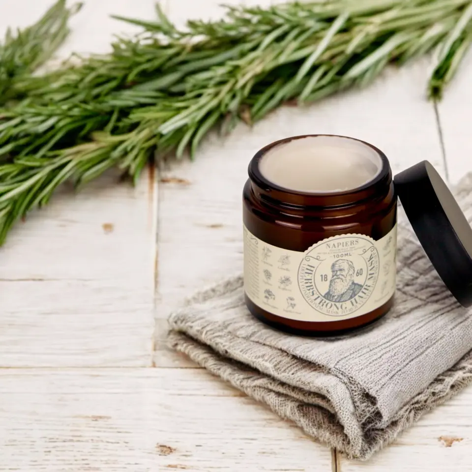 Brown jar of cream on grey cloth, rosemary sprigs in background.