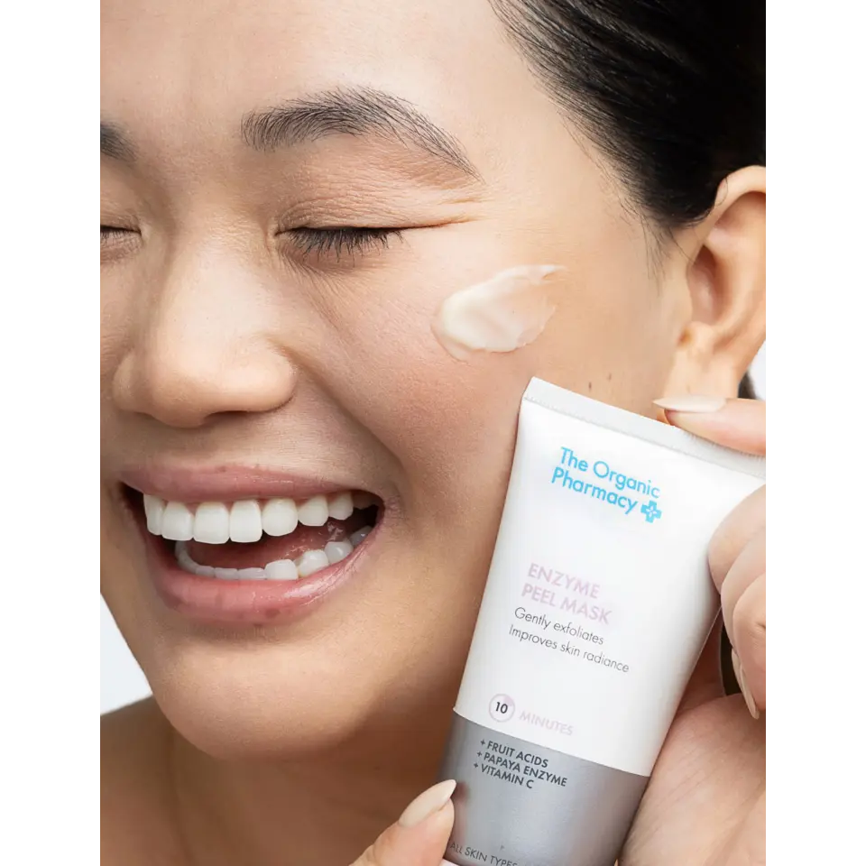 Smiling person applying The Organic Pharmacy Enzyme Peel Mask to cheek.