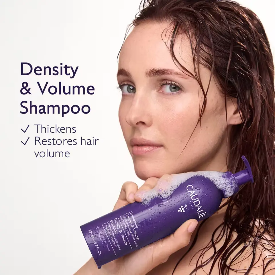 Woman holding Caudalie Density & Volume Shampoo bottle with wet hair. Text: Thickens, Restores hair volume.