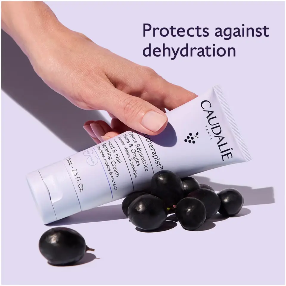 Hand holding Caudalie hand cream tube with grapes. Text: Protects against dehydration.