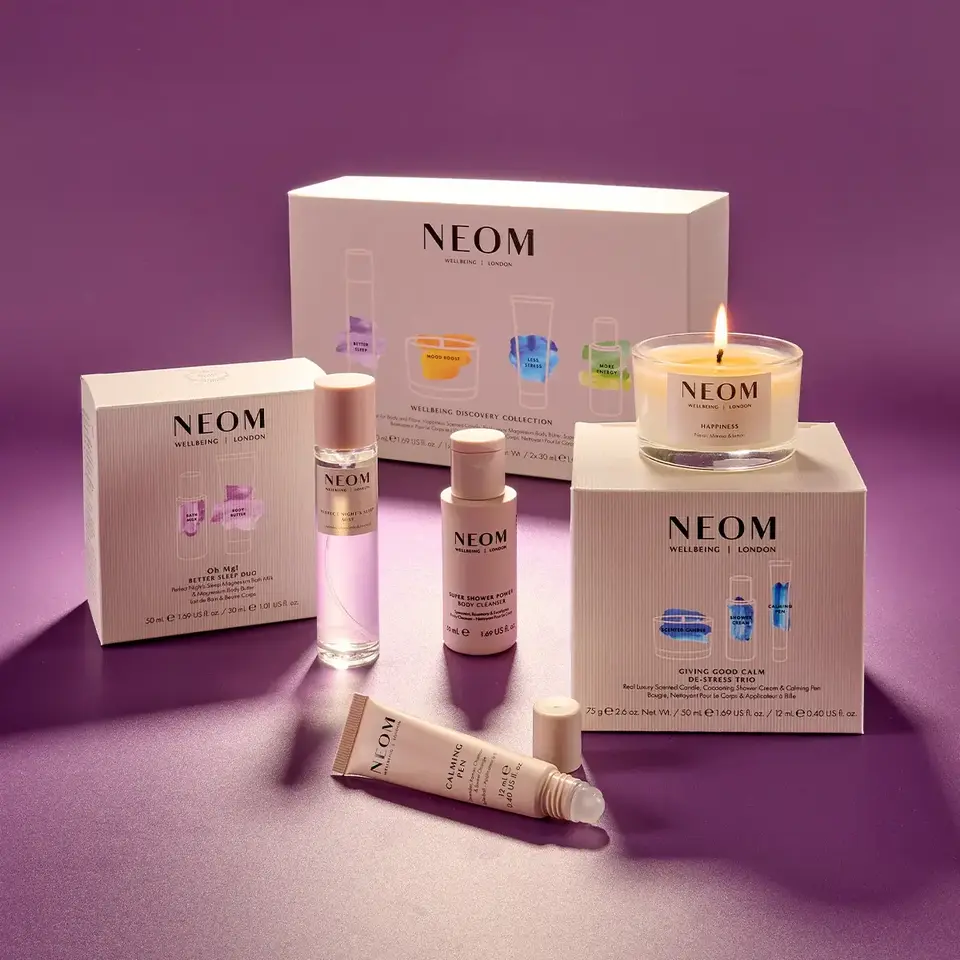 NEOM skincare and candle products on a purple surface, including boxed sets and a lit candle.