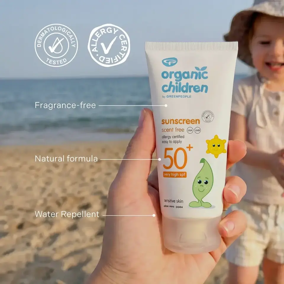 Hand holding Organic Children SPF 50+ sunscreen on a beach. Child in background.