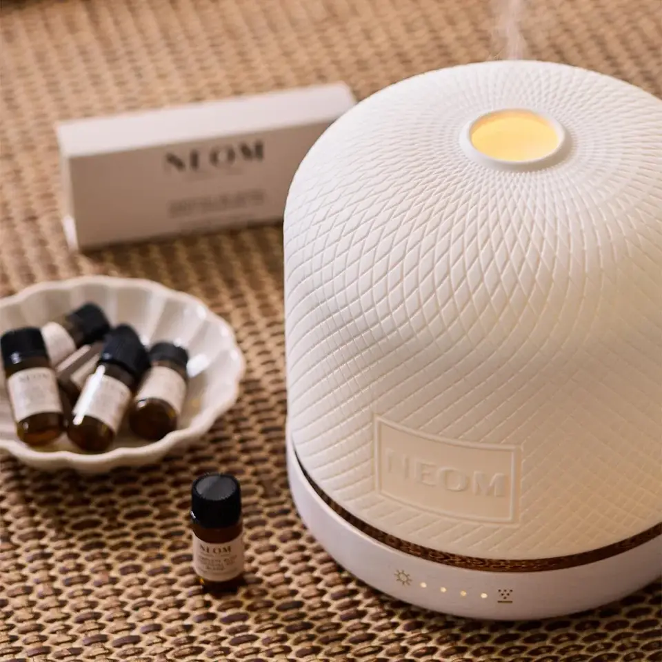 White textured diffuser with NEOM logo, surrounded by essential oil bottles on a woven mat.