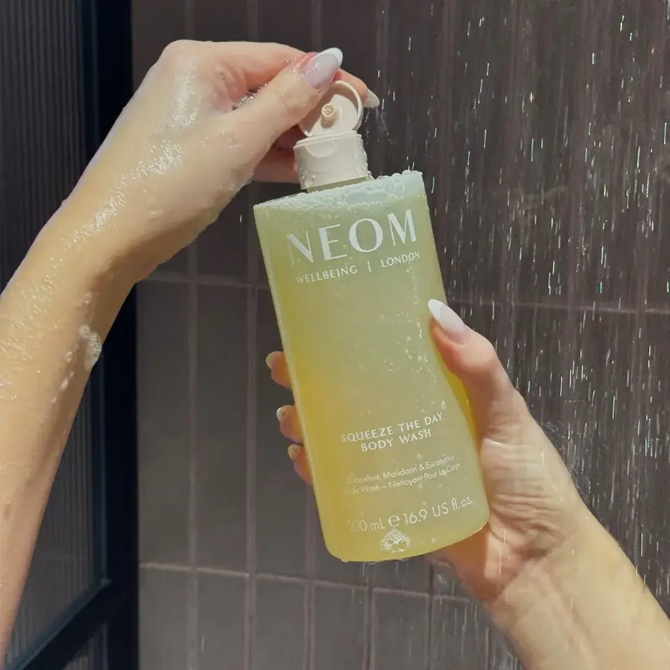 Hands holding Neom Squeeze the Day body wash in a shower.