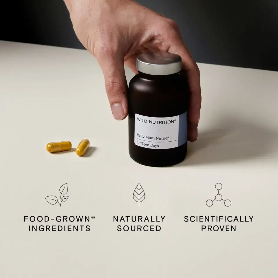 Hand holding a bottle of Wild Nutrition Daily Multi Nutrient for Teen Boys with two capsules nearby.