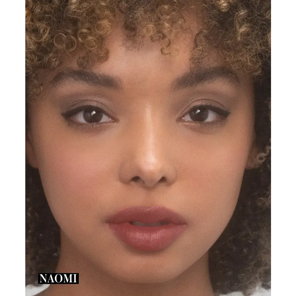 Close-up of a person with curly hair and natural makeup. Text: "NAOMI" in the corner.