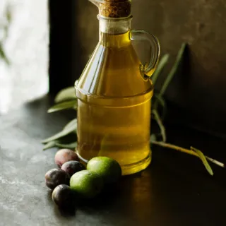 Glass bottle of olive oil with cork, surrounded by olives and leaves on a dark surface.
