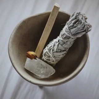 Bowl with sage bundle, palo santo stick, crystal, and dried rose on a white cloth background.
