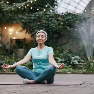 Woman meditating on a yoga mat in a greenhouse with plants and a water fountain.