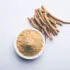 Bowl of ashwagandha powder next to dried roots on a white background.