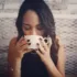 Woman with dark hair drinking from a white mug, seated against a brick wall.