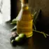 Glass bottle of olive oil with cork, surrounded by olives and leaves on a dark surface.