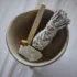 Bowl with sage bundle, palo santo stick, crystal, and dried rose on a white cloth background.