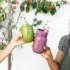 Two people holding green and purple smoothies in jars with straws, against a floral background.
