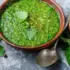Green nettle soup in a bowl with dried peppers, peppercorns, and fresh nettle leaves on a grey surface.