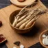 Ginseng roots in a wooden bowl on a bamboo mat, surrounded by herbs and dried ingredients.