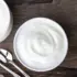 Two bowls of white cream with two spoons on a wooden surface.
