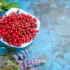 Red berries in a white bowl on a blue surface, surrounded by greenery and purple flowers.