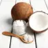 Whole and halved coconut with jar and spoon of coconut cream on white wood surface.