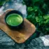 Bowl of green matcha on a wooden tray, placed on rocks with greenery in the background.