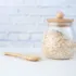 Glass jar filled with oats, wooden lid and spoon, against a white brick background.