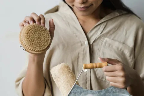 Woman holding a dry brush and loofah over a metal bucket.