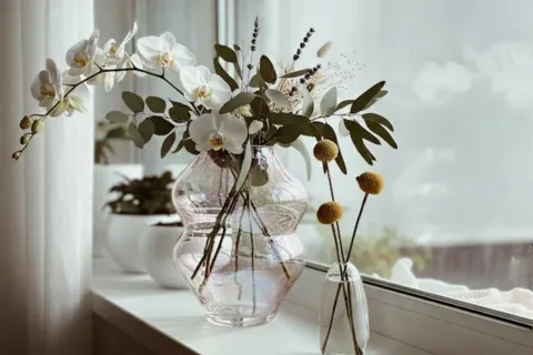 Orchids and foliage in a glass vase on a windowsill, with small yellow flowers in a separate vase.