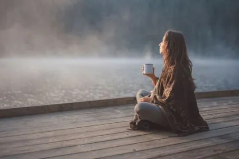 Person sitting on a wooden deck by a misty lake, holding a mug, facing the water.