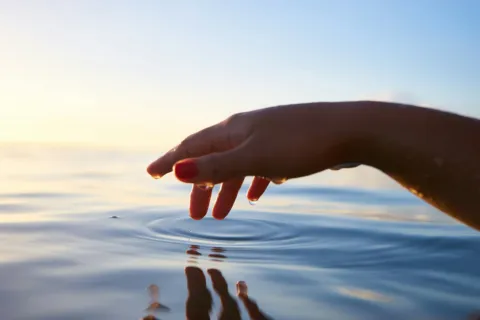 Hand with red nails touching calm water surface at sunset.