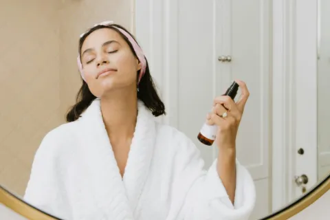 Person in a white robe using a spray bottle in front of a bathroom mirror.