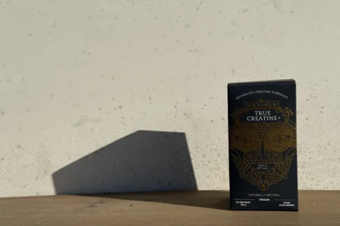 Box of True Creatine+ supplement on wooden surface, shadow cast on wall.