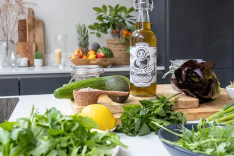Bottle of Honest Toil oil with fresh vegetables and herbs on a kitchen counter.