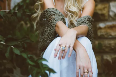 Woman with long hair wearing rings and a crochet shawl, sitting outdoors.
